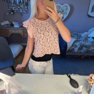 Ljusrosa spetsblus med volangärm - Supersöt ljusrosa blus i spets med korta volangärmar och rund hals. Blusen är croppad och har ett romantiskt mönster i spetsen som ger en feminin vibe. Perfekt att styla med jeans eller kjol för en trendig look.
