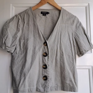 Ljusgrå blus från Soaked in Luxury - Supersnygg ljusgrå blus från Soaked in Luxury med korta puffärmar och djup v-ringning. Blusen har stora dekorativa knappar framtill och är tillverkad i ett lätt och luftigt tyg, perfekt för vår och sommar. Enkel att styla med både jeans och kjol.