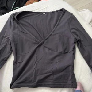 Mörkgrå omlottopp med v-ringning - Snygg mörkgrå omlottopp med djup v-ringning och långa ärmar. Toppens enkla design gör den lätt att matcha med olika stilar. Tillverkad i mjukt och stretchigt material som sitter bekvämt på kroppen.