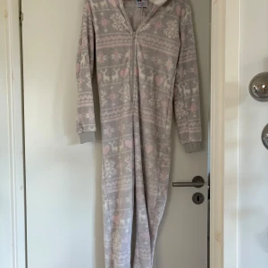 Mysig fleeceonesie med vintermönster - Supermjuk och varm onesie i ljusgrå fleece med vita och rosa vintermönster, hjärtan och renar. Har huva med fluffigt foder, dragkedja framtill och långa ärmar med muddar. Perfekt för kalla dagar när du vill chilla hemma. Aldrig använd. Strl S