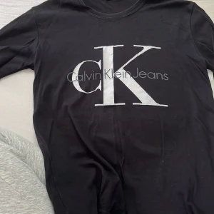Svart  t-shirt Calvin Klein - Svart  t-shirt från Calvin Klein Jeans med stort CK-tryck i silver på bröstet. Rund hals och klassisk passform, perfekt för dig som gillar stilrena logotoplagg. Materialet känns mjukt och skönt mot huden.