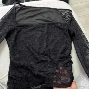 Svart spetsblus med meshdetalj - Snygg svart blus i spets med lång ärm och transparenta partier. Överdelen har en cool meshdetalj vid axlarna som ger en edgy vibe. Perfekt att styla med jeans eller kjol för en trendig look.