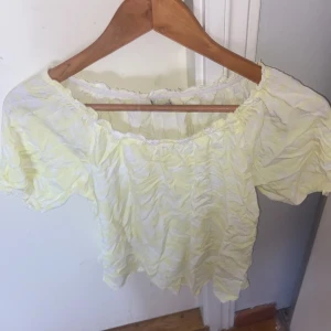Gul offshoulder blus med volang - Söt ljusgul blus med offshoulder-modell och korta puffärmar. Toppen har volangkant vid halsringningen och är lätt rynkad i tyget för en luftig känsla. Perfekt för vår och sommar när du vill ha något fräscht och ljust.