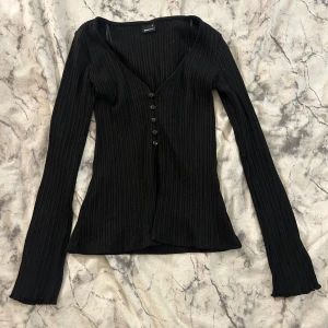 Svart ribbad långärmad topp - Svart ribbad topp från Gina tricot med v-ringning och knappar framtill halvägs. Långärmad modell i stretchigt material som sitter snyggt och markerar figuren. 