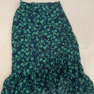 Blommig långkjol från Baum und Pferdgarten - Supersnygg långkjol från Baum und Pferdgarten med blått och grönt blommönster på svart botten. Kjolen har volang nertill och knappar längs sidan. Kjolen går precis ned till fötterna på mig som är 168cm. Pris går att diskuteras!