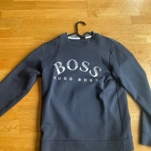 Mörkblå sweatshirt från Hugo Boss - Snygg mörkblå sweatshirt från Hugo Boss med stort BOSS-tryck i vitt och silver på bröstet. Tröjan har rund hals, långa ärmar och är gjord i mjuk bomull. Perfekt för dig som gillar stilrena och klassiska märkesplagg.