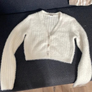 Vit fluffig kofta från Lindex - Supermjuk och fluffig vit kofta från Lindex med croppad passform. Koftan har ribbade muddar, v-ringning och tre glittriga knappar framtill. Perfekt för lager-på-lager och ger en mysig vibe till din outfit.