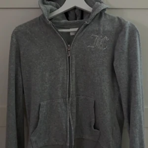 Grå Juicy Couture hoodie  - Grå hoodie från Juicy Couture med dragkedja, och huva.Strassdetaljer med JC-logga på bröstet och Juicy Couture-text på ryggen. Mjuk och skön.
