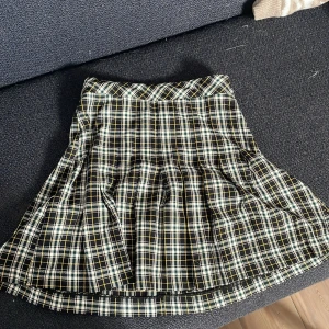 Rutig kortkjol från Lindex - Superfin rutig kortkjol från Lindex i svart, vit, gul och blå. Kjolen har klassiskt rutmönster, veckad design och stängs med knapp och dragkedja i sidan. Perfekt för en cool och trendig look.