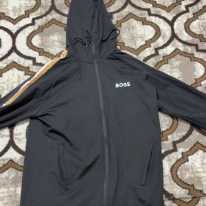 Svart kofta från Hugo Boss - Svart kofta från Hugo Boss med huva och hel dragkedja. Snygga beige och vita ränder längs ärmarna ger en sportig vibe. Jackan har två fickor framtill och justerbar snörning i huvan. Boss-logga på bröstet och detaljerad dragkedja.