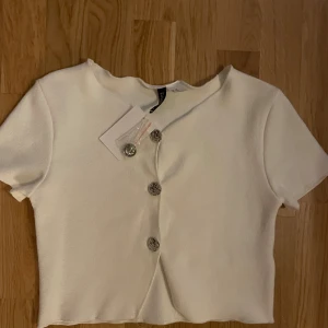 Vit kortärmad topp med silvriga knappar - Superfin vit topp från Daisy Street med korta ärmar och croppad passform. Toppen har snygga silvriga knappar framtill som ger en cool detalj. Perfekt till jeans eller kjol för en clean look.