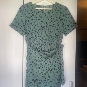 Grönprickig kort klänning från SHEIN - Superfin grön kortklänning från SHEIN med svarta prickar, rund hals och korta ärmar. Klänningen har ett knytband i midjan för extra snygg passform. Perfekt för dig som gillar lekfulla mönster och vill sticka ut lite extra. Någon fläck, se bild. Inget som syns speciellt.
