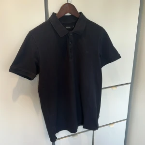 Svart pikétröja från Matinique - Svart pikétröja från Matinique i klassisk modell med krage och knappar framtill. Tröjan är i bomull och har en diskret broderad M-logga på bröstet. Perfekt för en clean och stilren look.