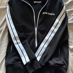 Palm Angels svart track jacket XL - Svart track jacket från Palm Angels i storlek XL med vita ränder längs ärmarna och dragkedja framtill. Tröjan har hög krage, ribbade muddar och logga på bröstet. Tillverkad i polyester för en sportig vibe och snygg passform.