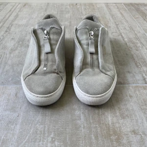 Grå sneakers från Axel Arigato - Snygga grå sneakers från Axel Arigato med mocka på ovansidan och detaljer i ljusgrått skinn vid hälen. Skorna har en unik dragkedja framtill istället för klassisk snörning och en vit platt sula. Perfekt för dig som gillar stilrena och moderna sneakers.