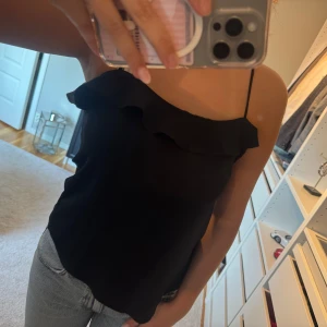 Svart volang-linnetopp med smala band - En svart linne-topp med smala axelband och snygg volangdetalj längs överdelen. 