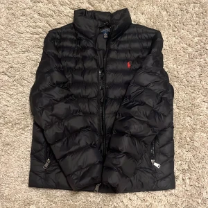 Svart pufferjacka från Polo Ralph Lauren - Säljer en svart pufferjacka från Polo. Jackan har quiltad design, dragkedja framtill och två sidofickor med dragkedja. Perfekt för dig som vill ha en stilren och varm jacka med snygg passform.  Jackan är storlek L/G 14-16 och jackan är inga defekter