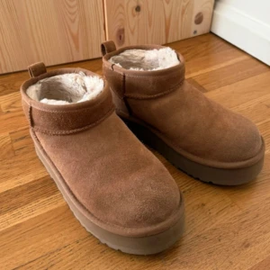Bruna UGG boots med platt sula - Säljer ett par bruna UGG boots med låg skaft och platt sula. Utsidan är i mjuk mocka och insidan är fodrad med fluffig päls för extra värme. Skorna har rund tå och en enkel, stilren design som är supermysig för kalla dagar. Har en del slitage i tyget och även tecken på användning i tyget inuti, köpt begagnat 
