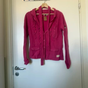 Rosa stickad kofta med volangdetaljer - Säljer en rosa stickad kofta från Odd Molly med långa ärmar, volangdetaljer framtill och knytband i midjan. Koftan har två fickor och broderad logga på ärmen samt siffran 506 broderad i nacken. Perfekt för dig som gillar färg och unika detaljer! Storlek 2 som enligt deras storleksguide motsvarar en storlek M.  Mer använd, noppror finns då materialet är ull och nylon. 