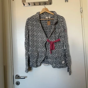 Mönstrad kofta med rosa knytband - Säljer en snygg kofta från Odd Molly i blått och vitt med grafiskt mönster. Koftan har ett rosa knytband i midjan, långa ärmar och mjukt stickat material. Perfekt för dig som gillar unika detaljer och vill ha något extra till garderoben. Storlek 2 som enligt deras storleksguide motsvarar en storlek M.  Fint skick, men har små tecken på användning. 