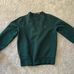 Grön crewneck från Polo Ralph Lauren - Mörkgrön crewneck-tröja från Polo Ralph Lauren med klassisk röd broderad logga på bröstet. Tröjan har rund halsringning, långa ärmar och är gjord i mjuk bomull. Perfekt för en chill och stilren look.