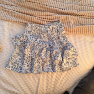 Vitblå blommig volangkjol - Supersöt kort kjol i vitt med blått blommönster och volanger. Kjolen har dragkedja i sidan och är perfekt för sommarens alla tillfällen. Lätt och luftig känsla, passar dig som gillar romantisk och fräsch stil.