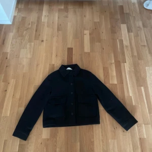 Svart höst jacka - Stilren svart overshirt med klassisk krage och stora fickor framtill. Jackan har knäppning med knappar hela vägen och långa ärmar med knappdetalj vid ärmslut. Perfekt för lager-på-lager och enkel att matcha till olika outfits.