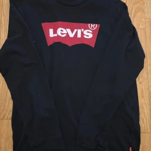 Svart långärmad Levi's tröja - Svart långärmad tröja från Levi's med klassiskt rött och vitt logotryck på bröstet. Tröjan har rund halsringning och är tillverkad i mjuk bomull, perfekt för en avslappnad och stilren look.
