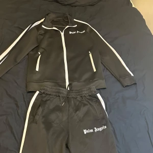 Svart tracksuit från Palm Angels - Svart tracksuit från Palm Angels med vita detaljer och dragkedja framtill. Jackan har vita ränder längs ärmarna och logga på bröstet. Snygg sportig look med ståkrage och två fickor med dragkedja. Perfekt för dig som gillar streetwear.