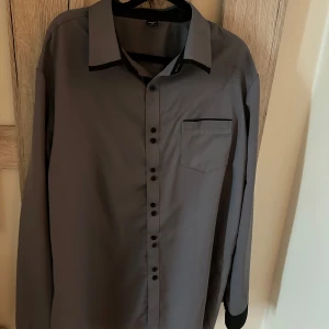 Grå skjorta med svarta detaljer - Stilren grå skjorta från Dressmann med svarta detaljer på krage, manschetter och knappslå. Skjortan har lång ärm, bröstficka och knappar framtill. Perfekt för dig som gillar en clean look med lite edge.