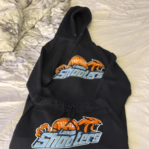 Svarta mjukisbyxor Trapstar Shooters - Svarta mjukisbyxor och hoodie från Trapstar med stort broderat motiv i orange, blått och vitt framtill. Byxorna har resår och snörning i midjan för skön passform. Perfekta för chill eller streetwear-stil. 