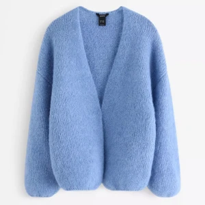 Blå stickad kofta från Lindex - Mysig blå stickad kofta från Lindex med oversized passform och lång ärm. Koftan har en djup v-ringning och är gjord i ett fluffigt, mjukt material som känns riktigt skönt mot huden. Perfekt att slänga på sig när det är kyligt ute.