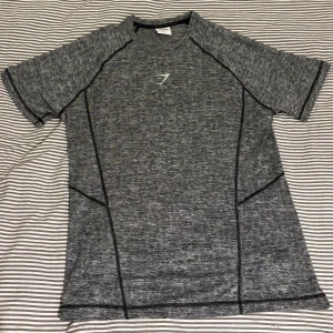 Grå Gymshark träningstshirt M - Säljer en gråmelerad t-shirt från Gymshark i storlek M. Perfekt för träning med sitt lätta och stretchiga material. T-shirten har korta ärmar, svarta sömdetaljer och en diskret Gymshark-logga på bröstet. T-shirten är aldrig använd.
