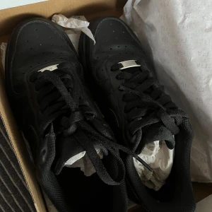 Svarta Nike Air Force 1 sneakers - Klassiska helsvarta Nike Air Force 1 sneakers i skinn med perforerad tå och svart snörning. perfekt för dig som gillar streetstyle. Ikonisk silverdetalj med 'AF1' på snörningen. Helt oanvända nypris är 1200kr