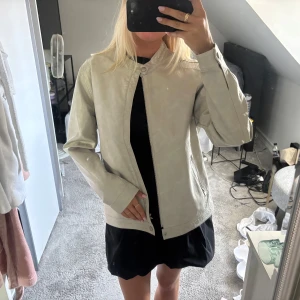 Beige skinnjacka med dragkedja - Snygg beige skinnjacka med minimalistisk design och raka linjer. Jackan har ståkrage med tryckknapp, dragkedja framtill och dragkedjeförsedda fickor. Perfekt för dig som gillar stilrena och tidlösa plagg. Långärmad och passar till många outfits.