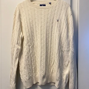 Vit kabelstickad tröja från GANT - Säljer en vit kabelstickad tröja från GANT i storlek XL. Tröjan har rund hals, ribbade muddar och ett broderat GANT-logo på bröstet. Perfekt för dig som gillar klassisk och clean stil. Materialet är mjukt och skönt, passar när du vill vara både chill och snygg.