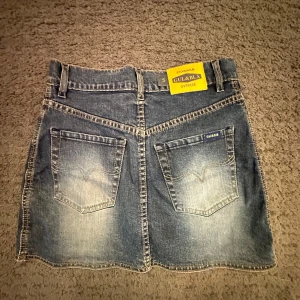 Blå jeanskjol från Gul & Blå - Snygg blå jeanskjol från Gul & Blå med klassisk femficksdesign, bälteshällor och dragkedja fram. Kjolen har slitna detaljer och tydliga sömmar, samt två stora bakfickor med logga. Perfekt för en chill och trendig look.