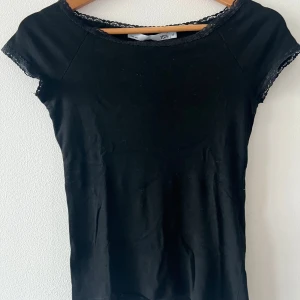 Svart topp med spets från 157 - En svart kortärmad topp från Lager 157 med fina spetsdetaljer längs halsringningen och ärmsluten. Toppens enkla design gör den lätt att matcha och den har en smickrande passform. Perfekt för dig som gillar stilrena plagg med en liten twist.