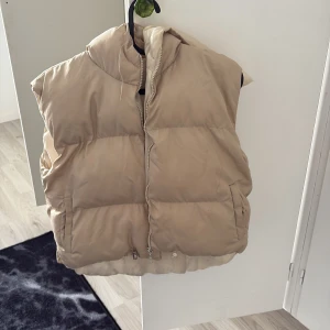 Beige dunväst med huva - Säljer en beige dunväst med huva och dragkedja framtill. Västen är quiltad och har två fickor på sidorna samt justerbar nederkant. Perfekt för lager-på-lager och chill höst- eller vinterstil.