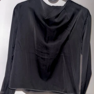 Svart långärmad blus med drapering - Stilren svart blus med lång ärm och snygg draperad urringning, från HM. Tunt, glansigt material som ger en elegant känsla. Perfekt att matcha med jeans eller kjol för en trendig look.