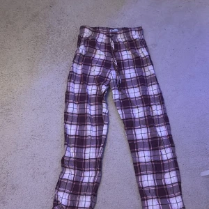 Rutig lila mjukisbyxa - Mysiga mjukisbyxor i lila och vitt rutigt mönster med dragsko i midjan. Perfekta för chill dagar hemma. Byxorna har rak passform och är gjorda i mjukt bomullstyg för extra komfort.