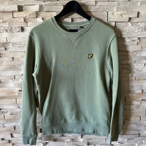 Grön sweatshirt från Lyle & Scott - Snygg ljusgrön sweatshirt från Lyle & Scott med klassisk rund hals och långärmad passform. Tröjan har det ikoniska gula fågelmärket broderat på bröstet och ribbade muddar vid ärmslut och nederkant. Perfekt för en chill och stilren look.