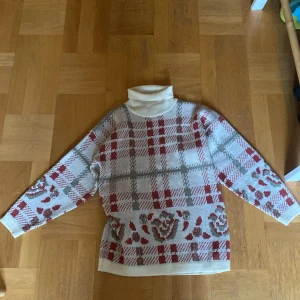 Rutig polotröja från Company i beige och rött - Säljer en beige polotröja från Company med rött och grönt rutmönster samt dekorativa detaljer nertill. Tröjan har lång ärm, hög krage och är gjord i 100% akryl. Perfekt för dig som gillar retrovibbar och vill sticka ut med ett unikt mönster. 