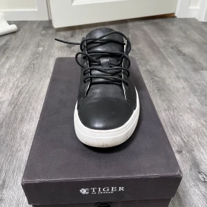 Svarta sneakers i skinn från Tiger of Sweden - Stilrena svarta sneakers i skinn från Tiger of Sweden med vit platt sula och klassisk snörning. Snygg minimalistisk design som passar perfekt till jeans,chinos eller till ett kostymset. Skorna har rund tå och är helsvarta med kontrasterande vit sula. Använda fåtal gånger. Nypris 2299. Originalbox tillkommer. 