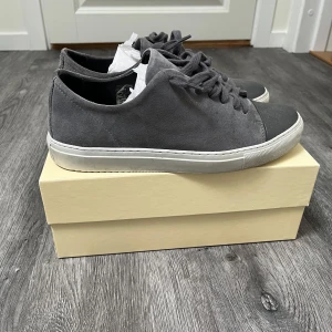 Grå sneakers från Axel Arigato mocka - Snygga grå sneakers från Axel Arigato i mocka med matchande grå snörning och vit sula. Skorna har en stilren design med rund tå och diskret logga i guld på sidan. Perfekta för dig som gillar minimalistisk och modern stil. Använda men har mycket kvar att ge. Nypris 1699. Originalbox tillkommer. 
