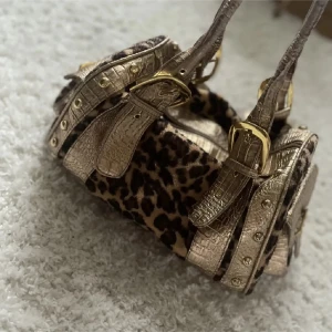 Leopardmönstrad handväska med guldiga detaljer - Fin handväska med leopardmönster och guldiga krokopräglade remmar och spännen. Perfekt för dig som vill sticka ut med en unik accessoar. Den enda defekten väskan har är att draggrejen på dragkedjan är lite trasig vilket gör det knepigare att öppna och stänga väskan men självklart funkar det fortfarande. Man kan även ha väskan som öppen hela tiden för att göra saker lättare men det ska gå att fixa tror jaggg. Köpt på plick av en tjej hsr andvänt några gånger 