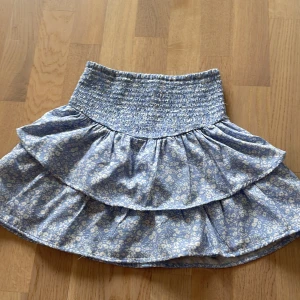 Blommig ljusblå volangkjol - Supersöt kort kjol med ljusblå botten och små vita blommor. Kjolen har bred smock i midjan och två lager volanger som ger en härlig och flowy känsla. Säljer pga är för liten. ❤️