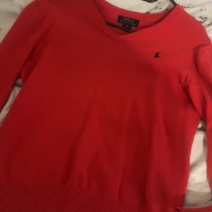 Röd v-ringad tröja från Polo Ralph Lauren - Säljer en snygg röd v-ringad tröja från Polo Ralph Lauren med den klassiska broderade loggan på bröstet. Tröjan har långa ärmar och ribbade muddar vid hals, ärmslut och nederkant. Perfekt för dig som gillar stilrena och tidlösa plagg.  Storlek M men passar mig som har XS/S