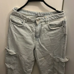 Ljusblå cargopants från Zara, strl 34 - Säljer ett par ljusblå cargopants från Zara i storlek 34. Byxorna har flera fickor, bland annat stora sidofickor med lock, klassisk jeanslook och raka ben. Materialet är jeans och passformen är loose för en chill vibe.