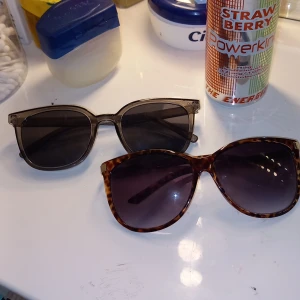 Två par solglasögon, leopard & svart - Säljer två par snygga solglasögon. Ett par har leopardmönstrad båge med guldiga detaljer och stora, runda glas. Det andra paret är helsvarta med klassisk fyrkantig form och mörka glas. Perfekta accessoarer för soliga dagar.